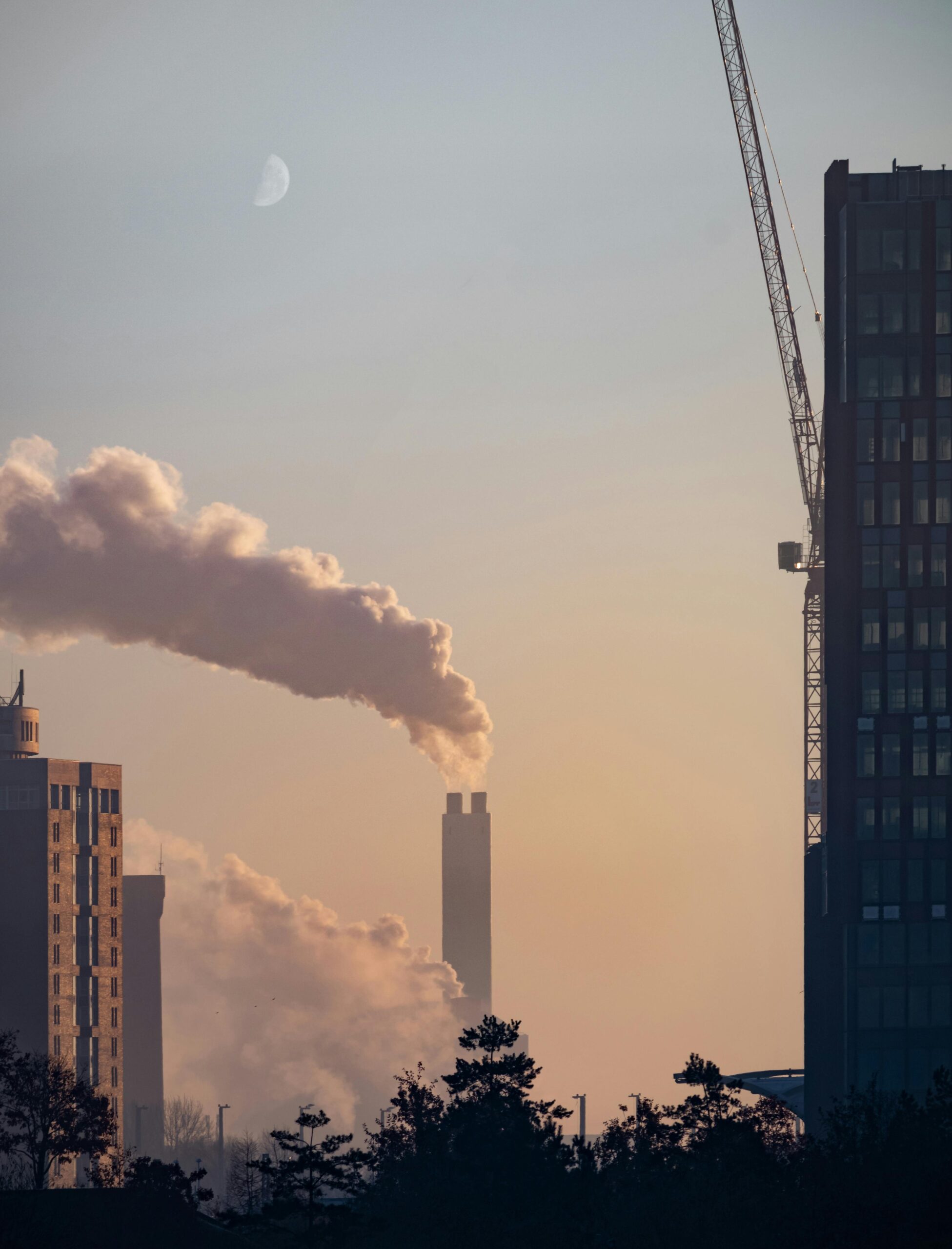 A cityscape featuring smokestacks emitting smoke, captured at sunrise with a visible moon.