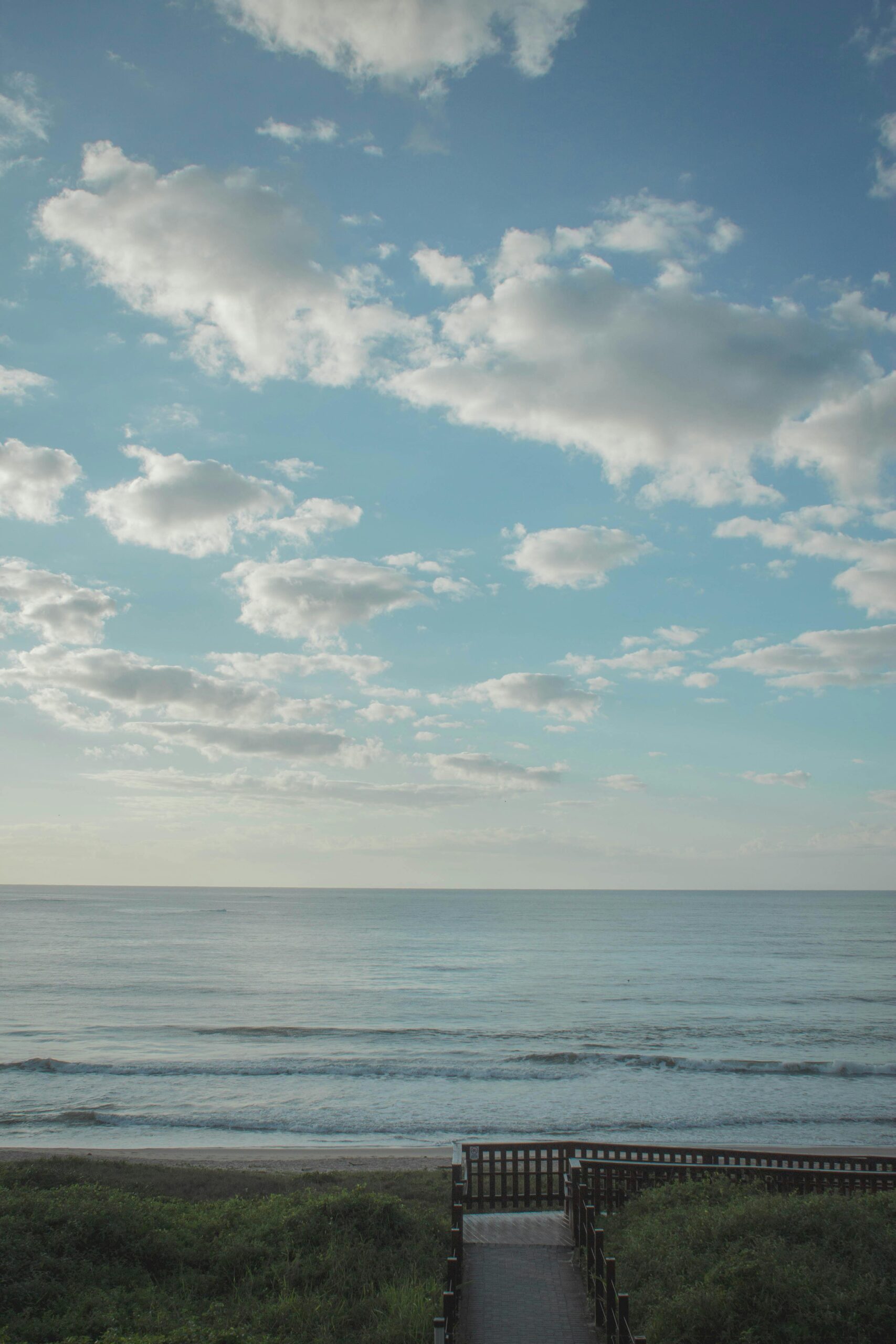 Serene view of a wooden pathway leading to the ocean with clouds in the blue sky.